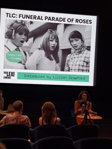 An audience watch a person speaking under a screen showing a still from Funeral Parade of Roses black and white photo