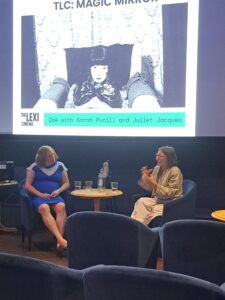 Two people speak on stage in front of a screen featuring a still of Magic Mirror, a black and white photo.