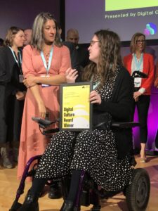 Photo from the ACE Digital Inclusion awards. A person who is a wheelchair user holds an award frame on a stage while talking to another person.