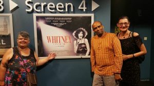 Friends stand around a poster of Whitney