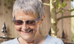 A older woman with short hair and sunglasses looks down, smiling, in a garden fence backdrop.