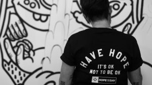 A person is stood in front of a white wall that has some artwork or graffiti on it. The person is wearing a top saying "Have hope, it's OK to not be OK".