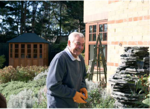 An older man in his garden smiling with gardening gloves on.