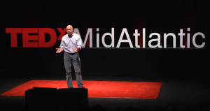 There is a man standing on a stage with "TEDX MidAtlantic" on the wall in the background.