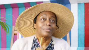 Screenshot from the shot film Gracie, an older Jamaican lady wearing a wide brimmed straw hat against a multi coloured striped background.