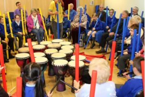 A group of young and older people in a workshop with bongos and brightly coloured tube instruments.