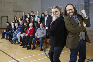 The Hairy Bikers stand in front of rows of people young and older in a school sports hall.