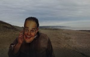 Still from the short film The Mask of a man walking along the coast wearing an Al Murray mask