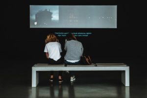 two people sitting watching a film in a gallery with subtitles