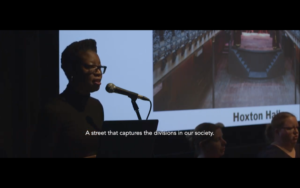 Karena speaks in front of a projected image of a theatre space. Caption reads: A street which highlights the divisions in our society."