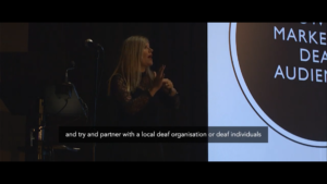 Nikki signs on stage. Caption reads: "try and partner with a local deaf organisation and deaf individuals"