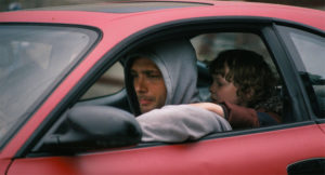 Still form Calm with Horses. Man with grey hoodie sitting in a red car with a child clinging to him from the seat behind him.