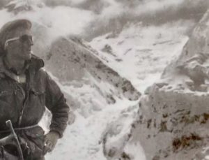 black and white image of a man in climbing gear and googles standing next to snowy mountains