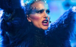 Natalie Portman in sparkly and feathered clothing and strong make-up including blue and purple streaks at the side of her face. Hr moth is open as she is singing and has a microphone attached to her.