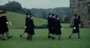 Still from an archive films about the British Red Cross Society Youth volunteers who are marching in uniform across green grass.