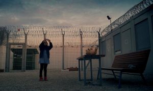 A still from Midnight Traveler. A young girl holds her phone up as if taking a selfie or a video in a detention centre setting with wire fencing.