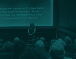 Green tinted photo of person standing in front of a cinema screen in front of an audience. There is text on the screen.