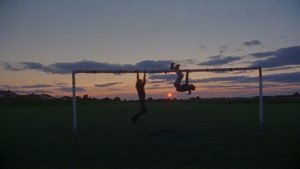Silhouettes of two people hanging from a goalpost in a field as the sun goes down.