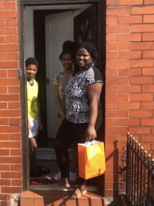 A family of mother and two children stand smiling in their home. The woman is holding a bright orange paper bag.