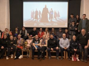 Group photo of the Young Carers film club at Spinners Mill. Two rows of people, mainly young people sit facing the camera smiling in front of a film screen.