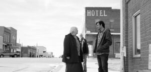 Black and White still from the film Nebraska. A younger man stands on the street on front of a Motel sign talking to an older lady while an older man stands behind them.