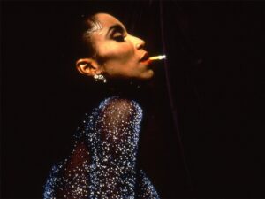 Still from Paris is Burning of a black person wearing a sparkly black outfit, red lipstick and chunky silver earrings performing with a cigarette in their mouth in front of a velvet curtain