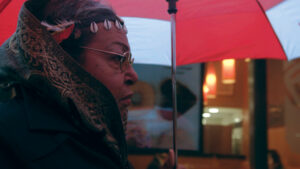 Still from the life and death of Marsha P Johnson. A side view of Marsha, a black transwoman wearing a dark coat and patterned scarf around her head and a shell headband, holding a red and white umbrella over her head.