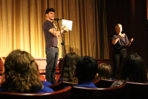 A person stands on a stage on front of a yellow lit curtain, holding and pointing to a script, a BSL Interpreter stands on the right hand side of them. Backs of heads of young people on cinema seats in the foreground.