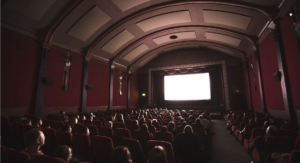 An audience sit in a large hall with red seats and walls in front of a cinema screen