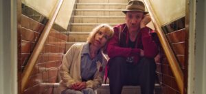 Sally Hawkins wears a blue dress and white cardigan, leans her head on David Thewlis' shoulder, as they sit on steps. David wears an old trilby hat, red shirt and jeans.