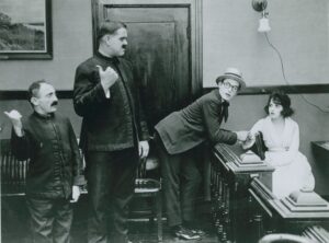 A short man, Sammy Brooks, Laurel and Hardy in a room with a woman , Bebe Daniels. Black and white silent film still photo.