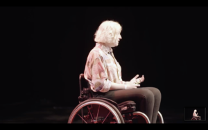 Amy has blond bobbed hair and faces to the right of the camera. She is gesticulating, wearing a pattered shirt, black trousers and sits in a wheelchair.