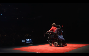 Judith has her back to us, she sits in a motorised wheelchair and wears a red t-shirt, she is positioned on a red carpet.