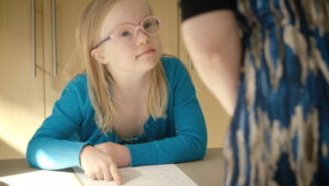 A girl with Down syndrome, long blonde hair, wearing glasses, and a blue top, points at a piece of paper on a desk. A woman is partially visible.