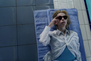 A blond woman lies on a towel on tiles by the side of a swimming pool. She wears a white shirt over a blue top, on a blue towel on blue tiles. She wears sunglasses.