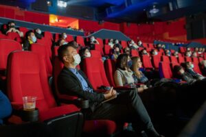 A cinema screen with people in it. The seats are red. The audience are attentively watching a film. There is social distancing in place and many seats are empty but the screen is still busy. People are wearing masks.