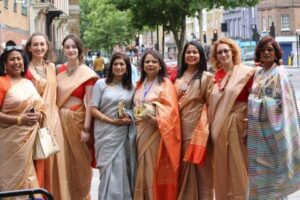Eight women of mixed heritages, spanning South Asian through White, wear orange and blue saris and smile to camera. The background is a city street with trees.