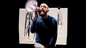 A balding white man with beard, wearing a blue jumper, appears to shout into a megaphone. Behind him is darkness with a white drawing featuring pen line marks.