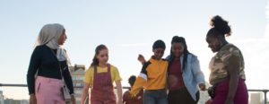 Still from Rocks - five young female-presenting people are on a rooftop dancing in the sunshine. They smile. They are a mix of ethnicities and sizes. and wear yellow, red, pink, black and camouflage, with one wearing a hijab.
