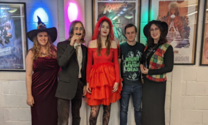 Photo of five young adults wearing halloween fancy dress standing in a row in front of a wall of movie posters.