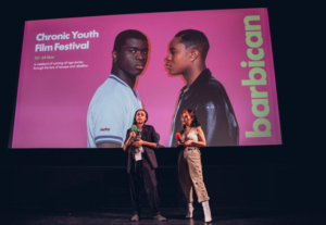 Photo from a cinema screening at Barbican. Two young people stand with microphones in front of a cinema screen which has a slide on it advertising Barbican "Chronic Youth Film Festival"