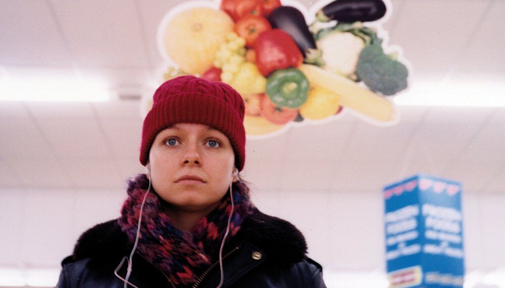 Still from the film Movern Callar, directed by Lynne Ramsey. A young woman played by Samantha Morton walks along a supermarket aisle wearing headphones, a red wool hat and red and black woolly scarf. She has a worried expression on her face.