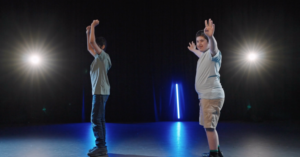 Screenshot from the dance film Moving Portraits. Two young boys dancing in a dark room with spotlights. They are standing sideways side by side with their hands in the air.