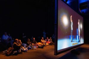 Photo of an audience on the floor behind the dance film Moving Portraits showing on a screen