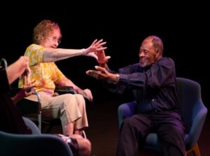 Photo of two people sitting on a stage communicating by tactile sign language.