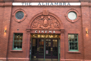 Photo of the outside of the Alhambra Cinema in Keswick. A grand looking red bricked building with glass doors, above which is a white "cinema sign" below old fashioned decorative detail in the architecture.