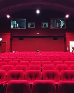 Red empty cinema seats, row by row, with a light above in an auditorium. A dark haired person is seated in the back row.