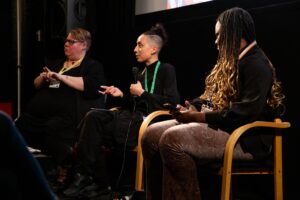 3 people, one who is BSL signing, sitting in front of a cinema screen talking at an industry event.