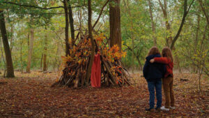Still from Petit Maman. Two children stand with their arms around each other looking at a den constructed from branches in an autumnal wood.