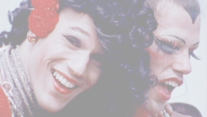 Still from the film Gay USA. A close up of two people smiling at a pride march in the 70's. They are both wearing vivid red lipstick. One person wears red flowers in their log black curly hair. The other person has big fluttery false eyelashes and a painted hairline.